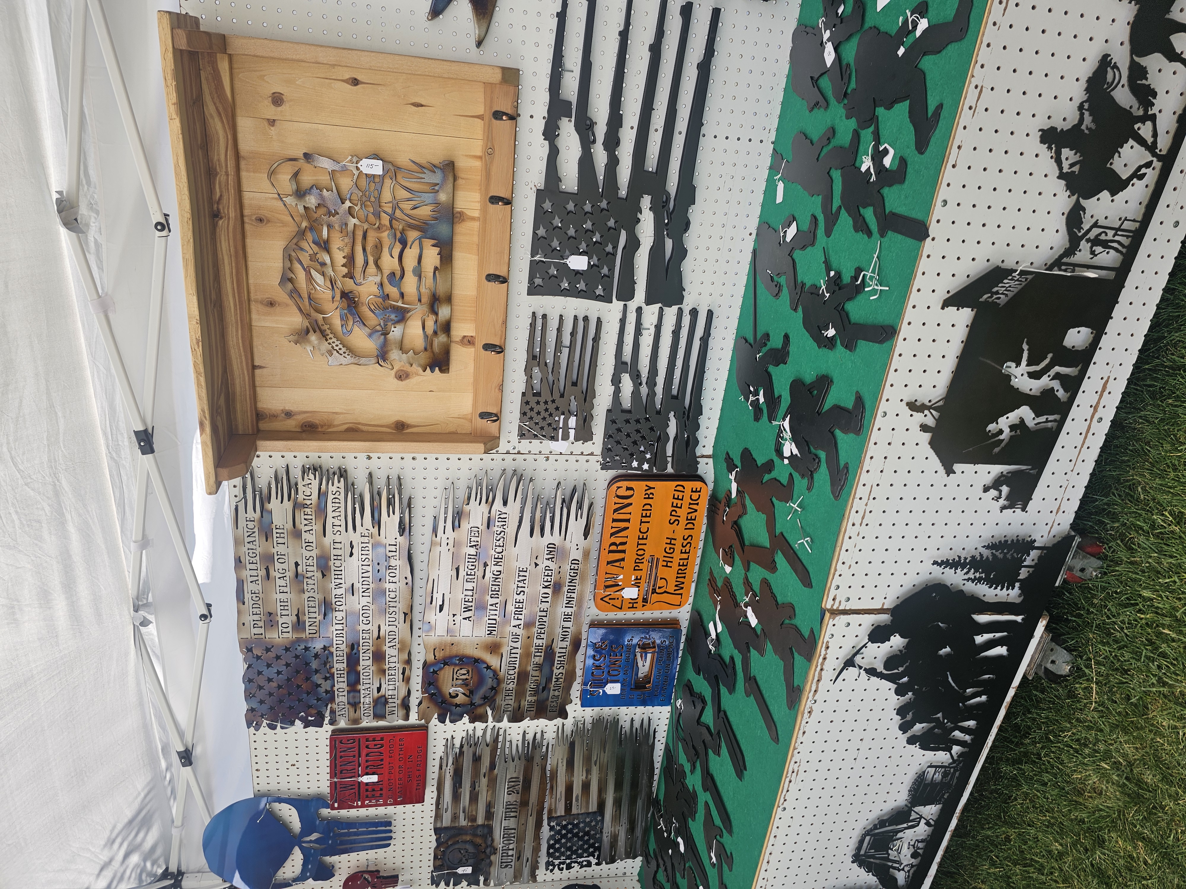 Market booth display of metal art with themes of hunting, fishing, and firearms, including a wooden coat rack with a fishing scene and flags made from rifle silhouettes.
