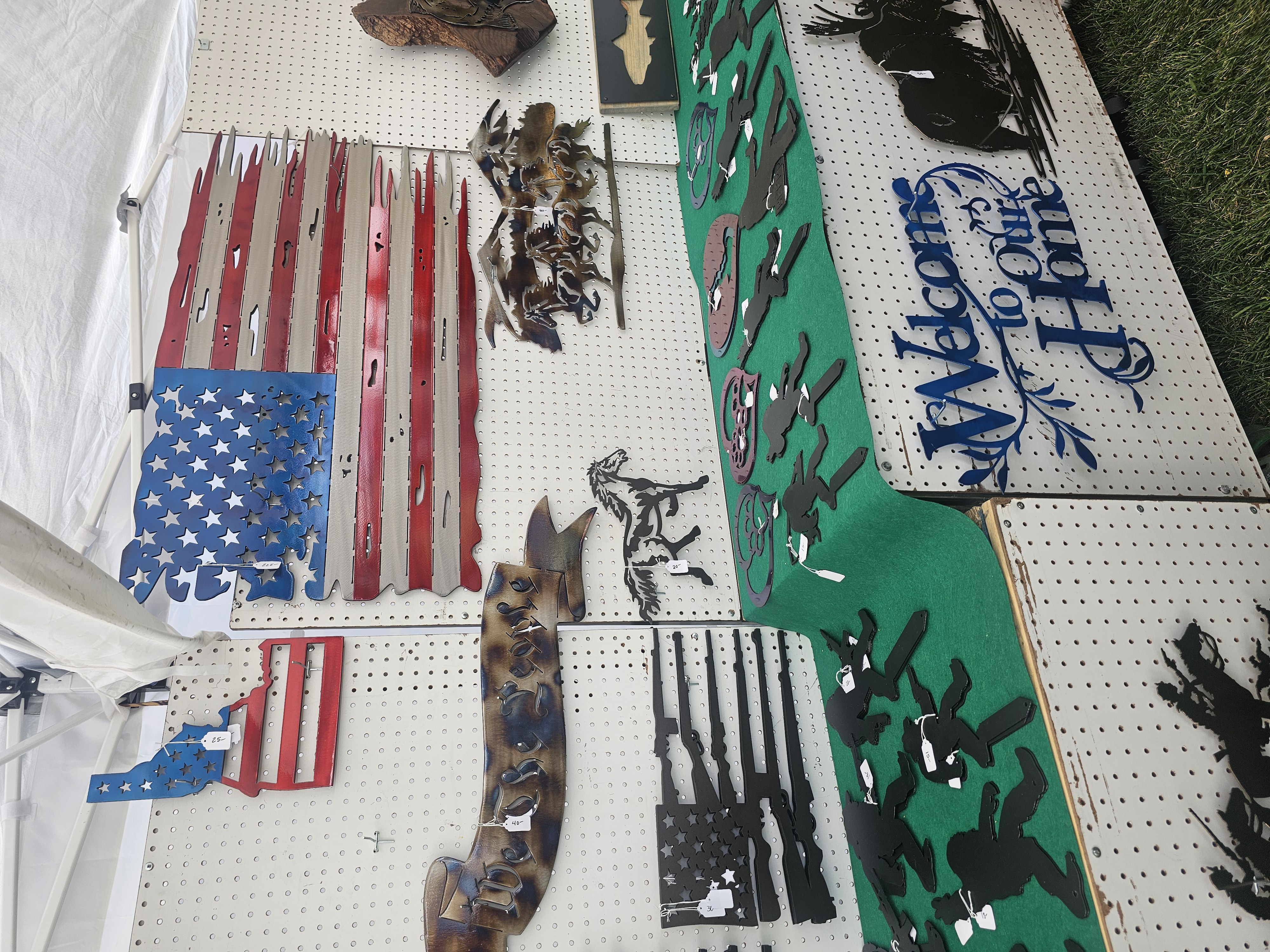 Market stall displaying home and western-themed metal art, featuring a 'Welcome to Our Home' sign, an Idaho state cutout, and a large tattered American flag.
